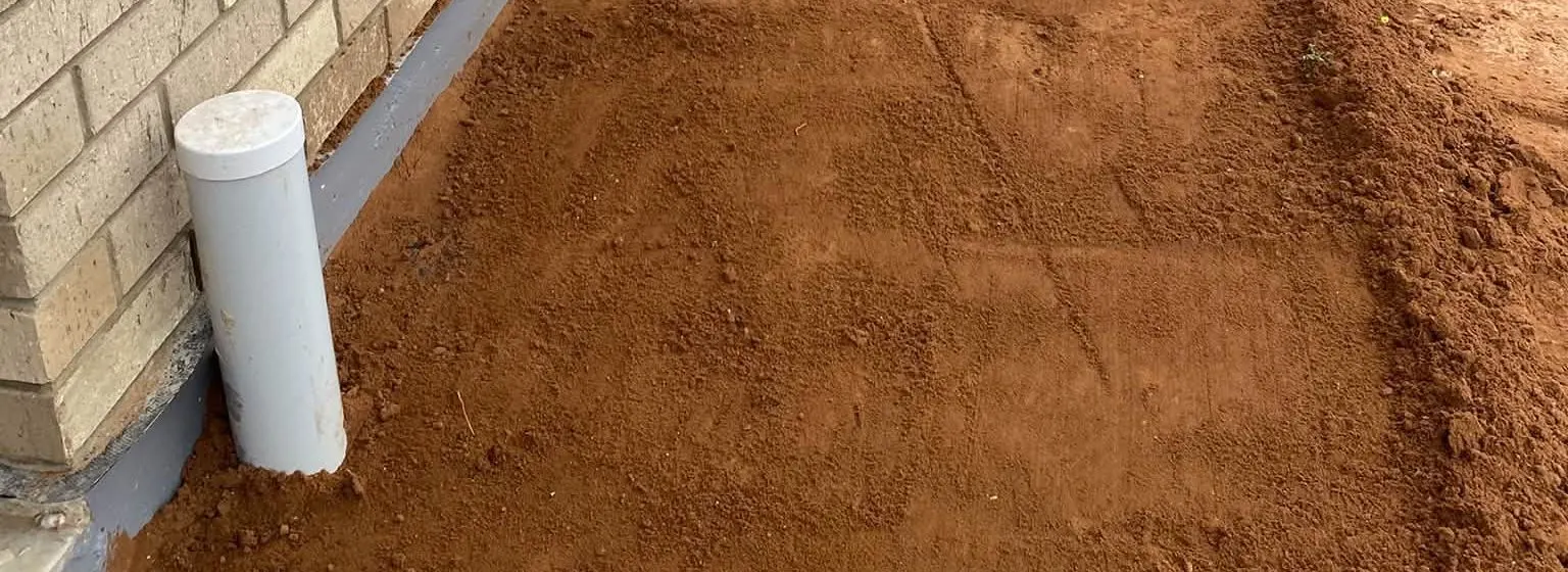 Red dirt levelled off along the base of a brick wall, a white pipe standing at the left