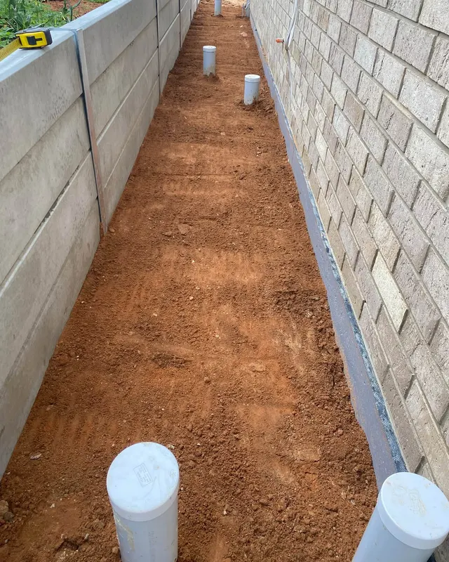 Prepped red dirt running along a concrete retaining wall with white pipes standing up